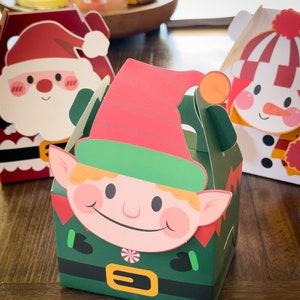 May include: Three Christmas-themed gift boxes with festive designs. The boxes feature Santa Claus, a snowman, and an elf. The boxes are made of paper and have a fold-over flap.