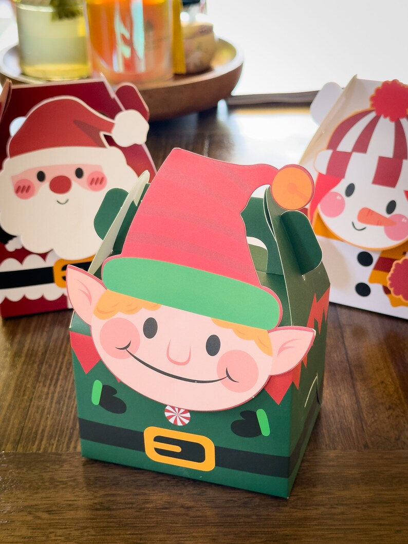 May include: Three Christmas-themed gift boxes with festive designs. The boxes feature Santa Claus, a snowman, and an elf. The boxes are made of paper and have a fold-over flap.