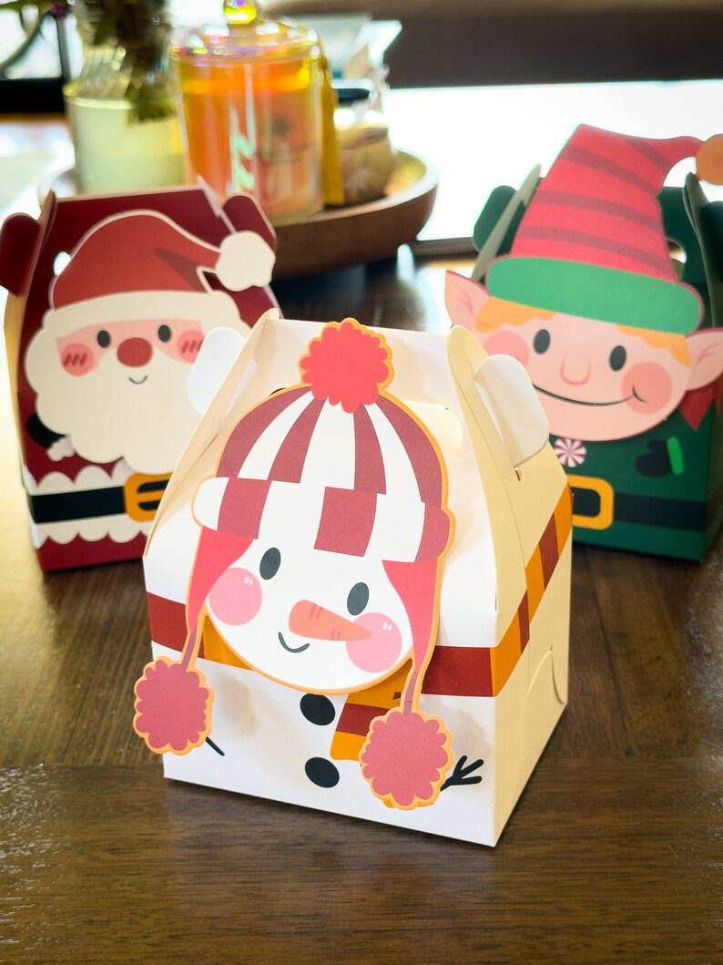 May include: Three Christmas-themed gift boxes with a snowman, Santa Claus, and an elf design. The boxes are made of cardboard and have a festive red, green, and white color scheme.