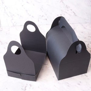 K&ouml;nnte beinhalten: Zwei schwarze Pappkartons mit Griffen. Die Kartons sind leer und haben ein schlichtes Design.