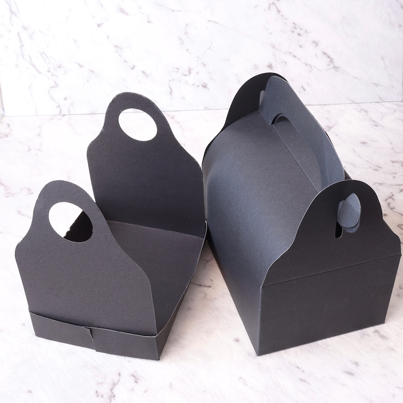 K&ouml;nnte beinhalten: Zwei schwarze Pappkartons mit Griffen. Die Kartons sind leer und haben ein schlichtes Design.