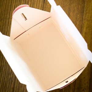 May include: A white cardboard takeout box with a pink interior. The box is empty and open, showing the inside.