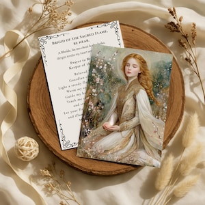Könnte beinhalten: Eine Karte mit dem Text "BRIGID OF THE SACRED FLAME, BE NEAR" und einer Illustration einer Frau mit rotem Haar und Heiligenschein. Die Karte liegt auf einem Holztablett mit Trockenblumen und einem dekorativen Ball.