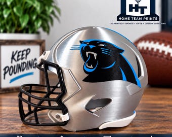 Puede incluir: Un casco de fútbol americano en miniatura de color plateado con un logotipo de pantera negro y azul en el lateral. El casco tiene una máscara facial negra y una correa de barbilla blanca. La superficie tiene un acabado metálico cepillado.