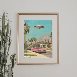 May include: A framed print of a pink car driving down a palm tree lined street with a flying saucer in the sky.