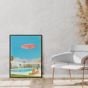 May include: A framed print of a mid-century modern home with a swimming pool and two people in bathing suits. A pink flying saucer is hovering over the house.