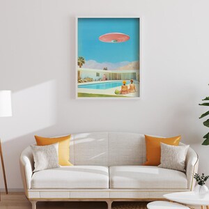 May include: A framed print of a mid-century modern home with a swimming pool and a pink flying saucer in the sky. Two people are sitting by the pool.