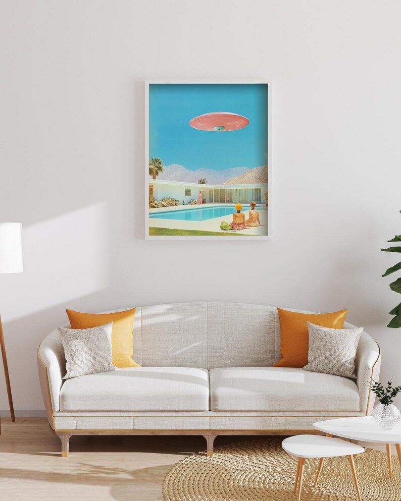 May include: A framed print of a mid-century modern home with a swimming pool and a pink flying saucer in the sky. Two people are sitting by the pool.