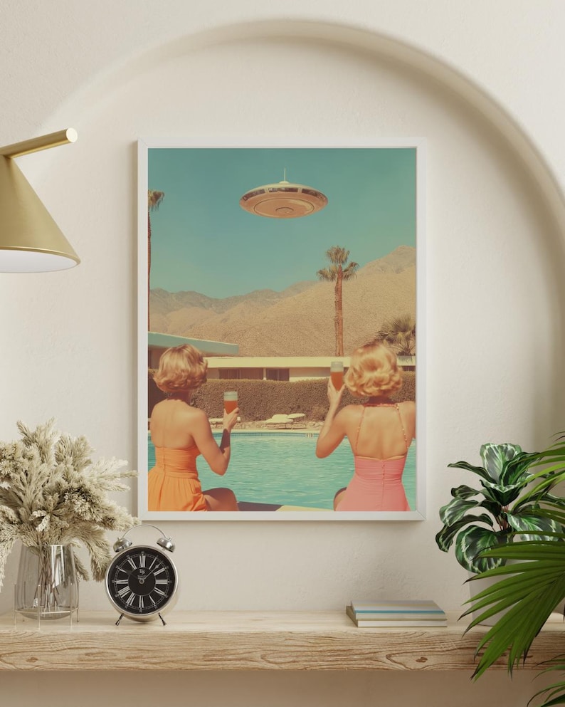May include: Two women in swimsuits sit by a pool, looking up at a flying saucer in the sky. They are each holding a glass of a light-colored beverage. The scene is set against a desert landscape with palm trees and a blue sky.