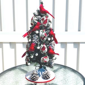 May include: A small Christmas tree decorated with red cardinal bird ornaments, white ball ornaments, and pinecone accents. The tree sits on a round base with two pinecones and a red trim. The tree is approximately 24 inches tall.