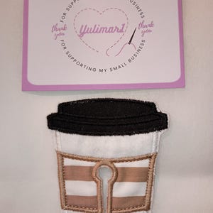 May include: A white and tan striped fabric patch shaped like a coffee cup with a black lid and two white plastic snaps at the bottom. Above the patch is a rectangular card with a purple border, a heart outline, a needle, and the text Yulimar1.