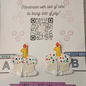 May include: Two white felt birthday cake appliques with colorful sprinkles and a single lit candle. The cakes are decorated with a white wavy line and a white border. The candles are yellow with a flame on top. The appliques are labeled "Style A" and "Style B". The text "Handmade with lots of care to bring lots of joy!" and "Made To Order!" are also visible.