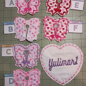 May include: Six different fabric bow designs with a heart pattern and a heart-shaped fabric with the name "Yulimarl" embroidered in purple glitter. The bows are pink, purple, and white with a heart pattern. The heart-shaped fabric is white with a pink border.
