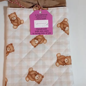 May include: A small, quilted sleeve with a repeating pattern of brown teddy bears on a cream and tan checkered background. The sleeve has a brown ribbon tie and a pink tag that reads "Quilted Sleeve Small".