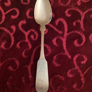 May include: A silver spoon with an ornate handle and a plain bowl. The back of the spoon is engraved with the initials "J.B.L."