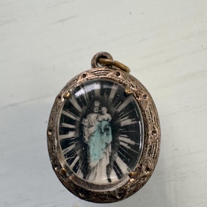 May include: An oval-shaped metal pendant featuring a central image of a figure holding a child, surrounded by a black and white sunburst pattern. The pendant has a textured, tarnished bronze-colored frame with a small loop at the top for hanging.
