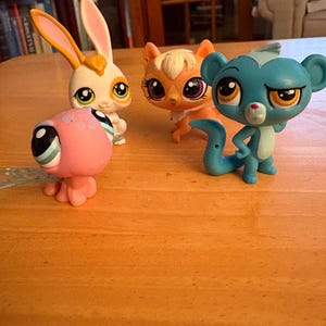 May include: Four colorful collectible animal figurines are arranged on a wooden surface. The figurines include a white bunny with orange ears, an orange cat, a pink creature, and a blue monkey. Each figure has large eyes and is designed in a cartoon style.