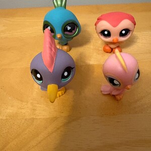 May include: Four colorful bird figurines with large eyes are arranged on a wooden surface. The birds are in shades of blue, red, purple, and pink, with yellow beaks and various crests. These collectible toys are designed with a cartoonish aesthetic.