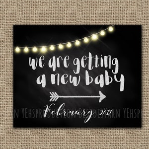 May include: Black chalkboard sign with white string lights, an arrow pointing to the right, and the text "We are getting a new baby February 2017".