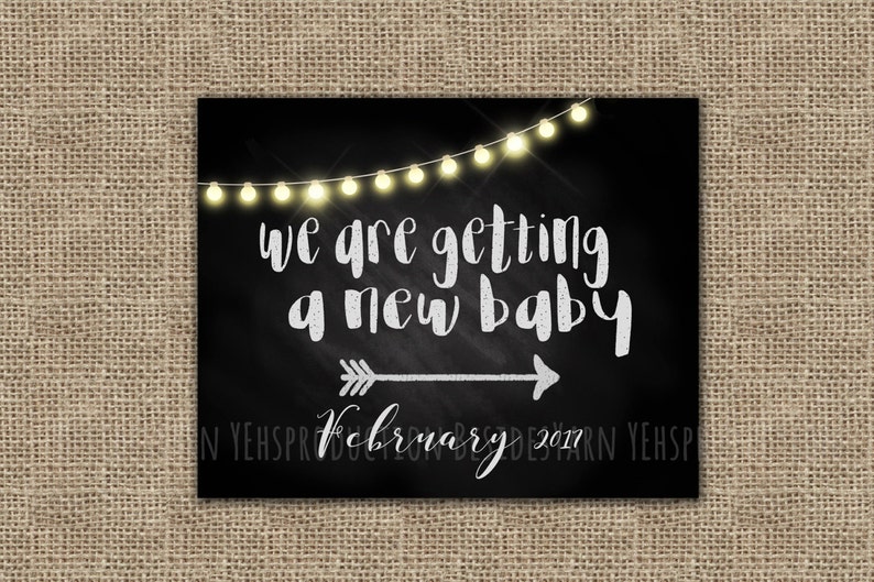 May include: Black chalkboard sign with white string lights, an arrow pointing to the right, and the text "We are getting a new baby February 2017".