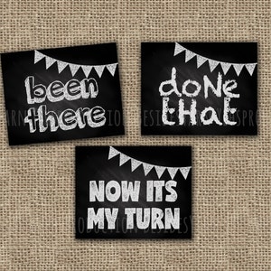 May include: Three black chalkboard signs with white chalk lettering and a string of white chalk flags hanging above each sign. The signs read "been there", "done that", and "now its my turn".