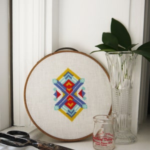 May include: A white embroidery hoop with a colourful geometric design stitched in the centre. The hoop is sitting on a windowsill with a pair of scissors and a small glass bottle with red text that reads "Standard Dairy, Alabama's Finest Milk".