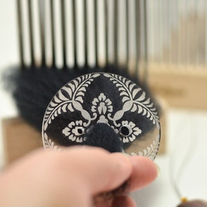 May include: A clear plastic disc with a white floral pattern and two small holes. The disc is used for felting wool.