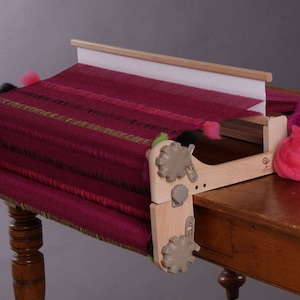 May include: A wooden loom with a burgundy and green woven fabric on it. The loom is set up on a wooden table with balls of pink yarn.