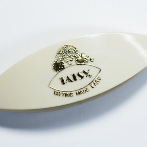 May include: An off-white, almond-shaped tatting shuttle featuring a gold-colored logo on the center. The logo depicts a stylized floral design above the text TATSY and the phrase TATTING MADE EASY printed in a clear, gold-colored font.
