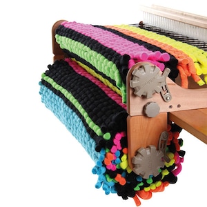 May include: A wooden loom with two colorful rugs rolled up on the side. The rugs are made with thick yarn and have a variety of colors, including black, green, blue, pink, yellow, and orange. The loom has a wooden frame and metal parts.