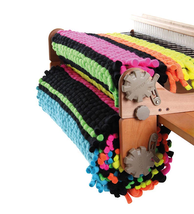 May include: A wooden loom with two colorful rugs rolled up on the side. The rugs are made with thick yarn and have a variety of colors, including black, green, blue, pink, yellow, and orange. The loom has a wooden frame and metal parts.