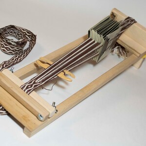 Puede incluir: Un telar de madera con una banda tejida marr&oacute;n y blanca. El telar tiene un marco rectangular con una serie de tarjetas y una lanzadera. La banda tejida tiene un dise&ntilde;o geom&eacute;trico repetitivo.