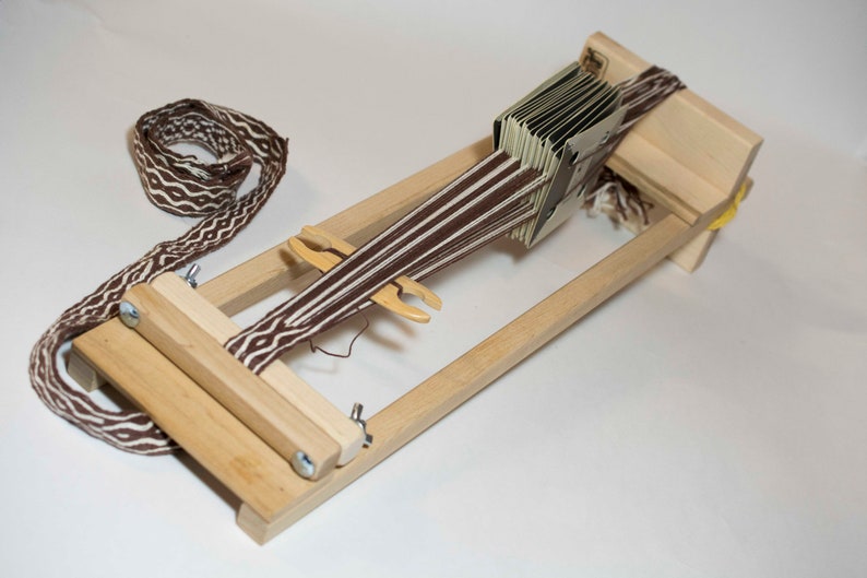 Puede incluir: Un telar de madera con una banda tejida marr&oacute;n y blanca. El telar tiene un marco rectangular con una serie de tarjetas y una lanzadera. La banda tejida tiene un dise&ntilde;o geom&eacute;trico repetitivo.