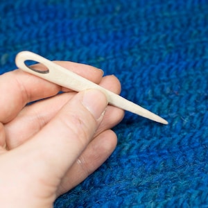 Puede incluir: Primer plano de una mano sosteniendo una aguja de tejer de madera con un peque&ntilde;o agujero en la parte superior. La aguja descansa sobre una tela de punto azul.