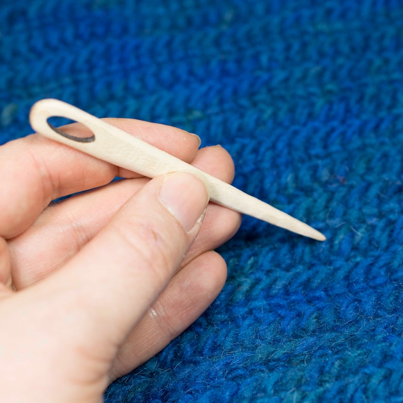 Puede incluir: Primer plano de una mano sosteniendo una aguja de tejer de madera con un peque&ntilde;o agujero en la parte superior. La aguja descansa sobre una tela de punto azul.