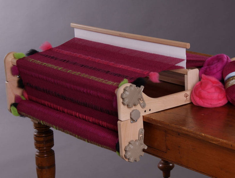 May include: A wooden loom with a burgundy and green woven fabric on it. The loom is set up on a wooden table with balls of pink yarn.