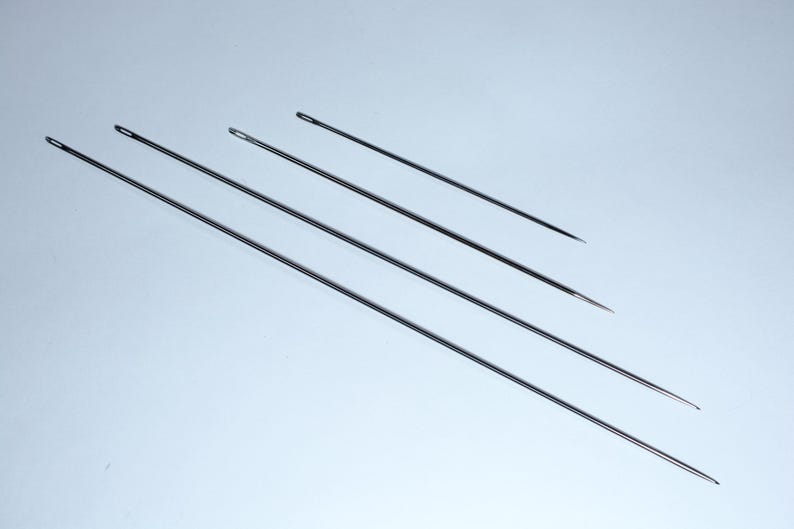 May include: Four silver sewing needles of varying lengths, arranged diagonally on a white background.