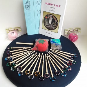 May include: A bobbin lace kit with a blue cushion, wooden bobbins, a red pincushion, a needle threader, and two books titled "Bobbin Lace Without a Teacher" by Betty Alderson.