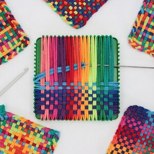 May include: A close-up of a loom with colorful yarn being woven into a square coaster. The loom is green and has a needle and crochet hook nearby. There are four finished coasters in the background, all with a rainbow pattern.