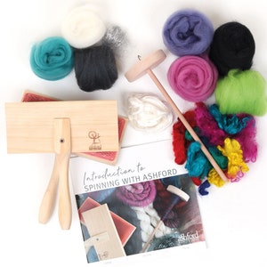 May include: A spinning wheel kit with a wooden spindle, a wooden carding brush, and a booklet titled "Introduction to Spinning with Ashford". The kit includes various colors of wool roving, including white, gray, teal, purple, pink, black, and green.