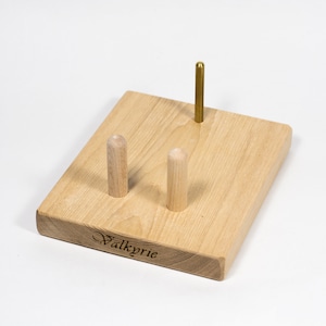 May include: Wooden stand with three pegs for holding yarn or thread. The stand is light brown and has the word "Valkyrie" engraved on the bottom.