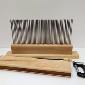 May include: A wooden wool carding set with a metal comb, a wooden handle, and a clamp. The comb has a wooden base and a row of metal teeth. The handle is made of wood and has a metal pin. The clamp is made of metal and has a black handle.