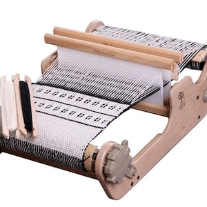 May include: A wooden table loom with a white and black woven fabric on it. The loom has two wooden shuttles and two crank handles. There are three spools of yarn on the loom, one white and two black.