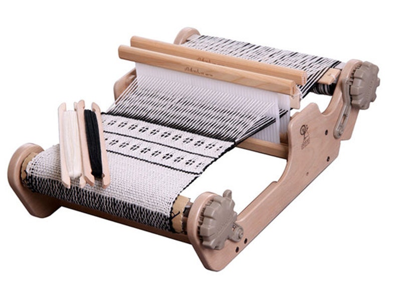 May include: A wooden table loom with a white and black woven fabric on it. The loom has two wooden shuttles and two crank handles. There are three spools of yarn on the loom, one white and two black.