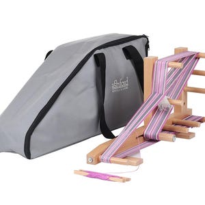 May include: A light-colored wooden warping board with multiple pegs, wrapped in a striped pink, purple, and white woven fabric. A separate small wooden shuttle with pink yarn sits in front, alongside a grey carrying bag with the Ashford logo.