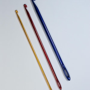 May include: Three crochet hooks in gold, red, and blue. The hooks are made of metal and have a smooth, shiny finish. The hooks are different sizes, with the gold hook being the smallest and the blue hook being the largest. The hooks are arranged diagonally.