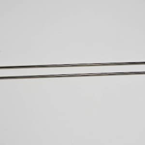 May include: Two silver sewing needles, one slightly longer than the other, are lying parallel on a white surface.
