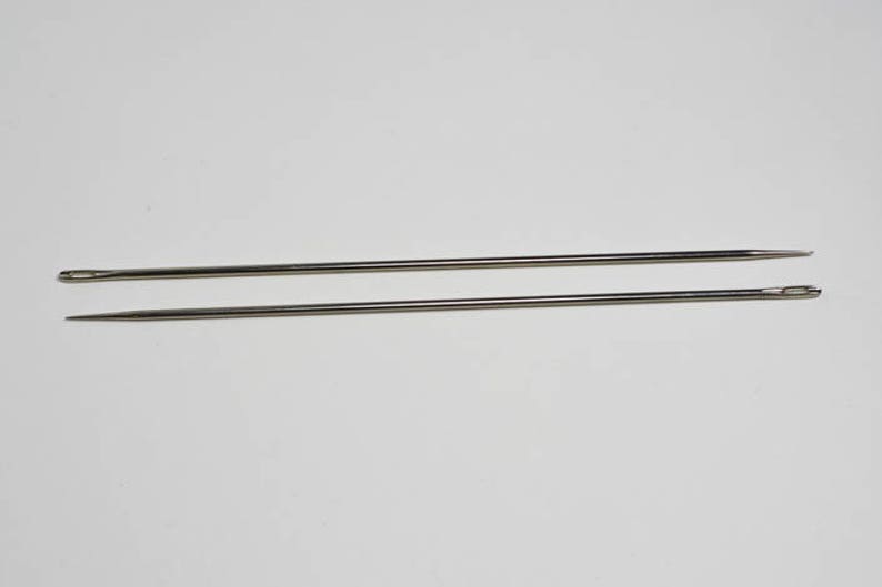 May include: Two silver sewing needles, one slightly longer than the other, are lying parallel on a white surface.