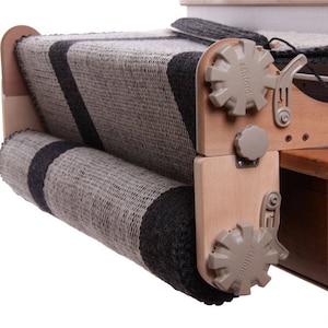 May include: A close-up of a wooden loom with a gray and black striped fabric being woven. The loom has two tension knobs labeled "Ashford" and "Pro-Baber".