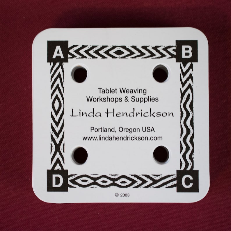 Puede incluir: Una herramienta de tejido de tableta cuadrada blanca con cuatro agujeros y un borde geom&eacute;trico en blanco y negro. El texto de la herramienta dice "Tablet Weaving Workshops & Supplies" y "Linda Hendrickson, Portland, Oregon USA".